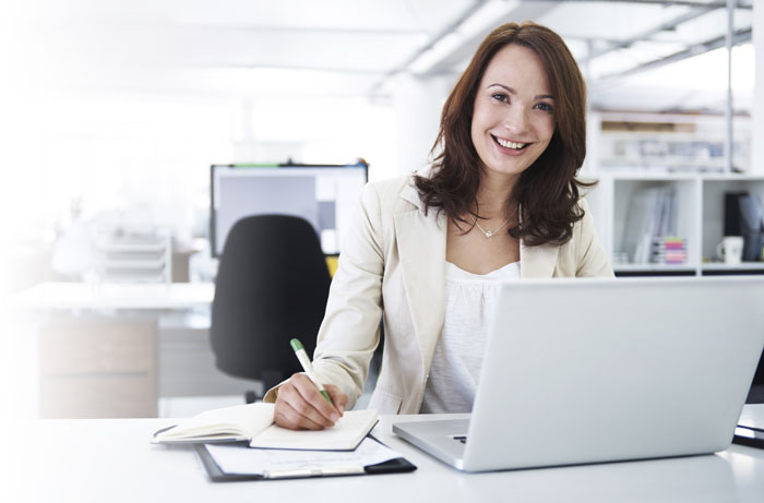A smiling, young businesswoman writing down notes and working on her laptop